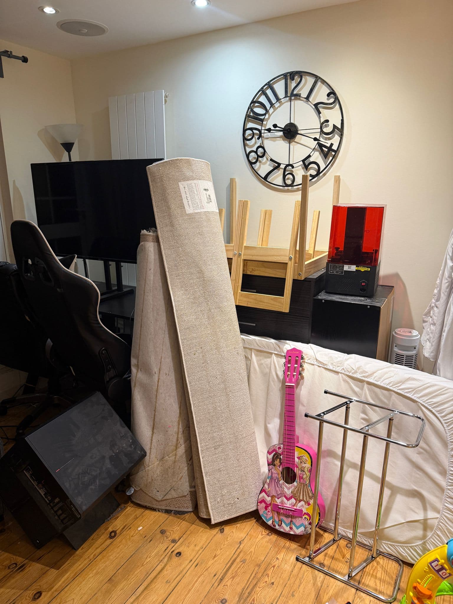 Cluttered room with rolled rugs, a pink toy guitar, electronics, and a large wall clock.